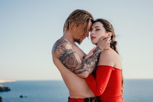 Portrait of tattooed man and sensual young brunette woman in red dress, happily dancing outdoors isolated on blurry sea background with natural bokeh in soft warm sunset backlight. Selective focus