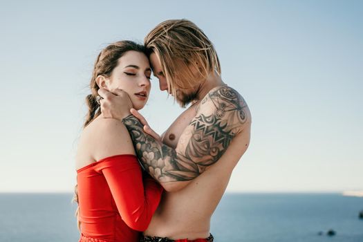 Portrait of tattooed man and sensual young brunette woman in red dress, happily dancing outdoors isolated on blurry sea background with natural bokeh in soft warm sunset backlight. Selective focus