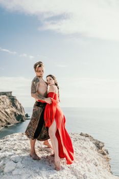 Portrait of tattooed man and sensual young brunette woman in red dress, happily dancing outdoors isolated on blurry sea background with natural bokeh in soft warm sunset backlight. Selective focus