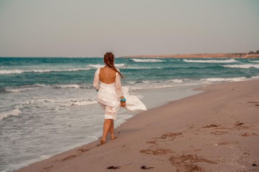 Model in boho style in a white long dress and silver jewelry on the beach. Her hair is braided, and there are many bracelets on her arms