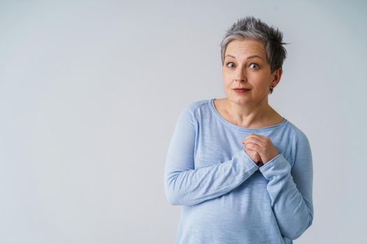 Amazed mature woman in 50s with grey hair posing with hands up next to heart and copy space on left isolated on white background. Copy space and place for product placement. Aged beauty. Toned image.