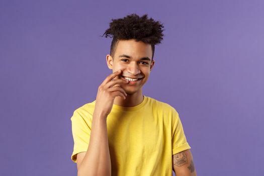 Close-up portrait of lovely young hispanic gay man with dreads, tattoos, touching lip sensually and flirty smiling, checking out someone really cute and handsome, standing purple background.