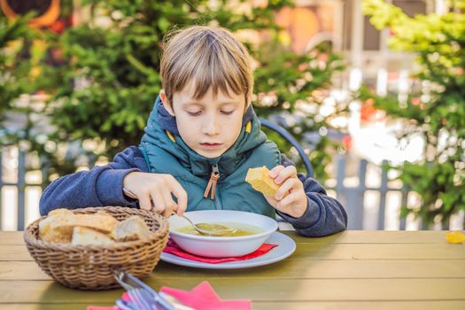 Adorable little school boy eating vegetable soup outdoor. Blond child in domestic kitchen or in school canteen. Cute kid and healthy food, organic vegan soup with potato, tomato and vegetables.