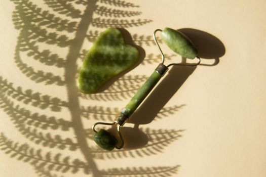 Jade Gua sha scraper and face roller massager on a cork round stand with a monstera leaf. Hard light, shadows, the concept self-care. Facial care. Zero waste. Lifting and toning treatment at home.