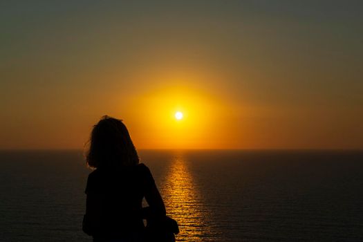 Silhouette of a woman at orange sunset on a cliff overlooking the sea at Palaiokastro castle of ancient Pylos. Greece.