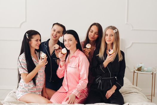 Stock photo of pretty bridesmaids in pyjamas holding delicious handmade cupcakes in front of their lips looking at camera. Celebrating hen party together. Best friends.