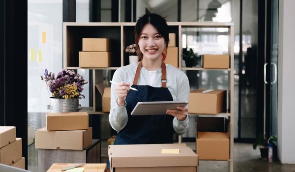 Asian woman writing customer's shipping information on parcel box, she owns an online store, she ships products to customers through a private courier company. Online selling concept..