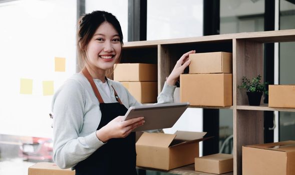 Young beautiful happy asian business woman owner of SME online using laptop receive order from customer with parcel box packaging at her startup home office, online business seller and delivery.