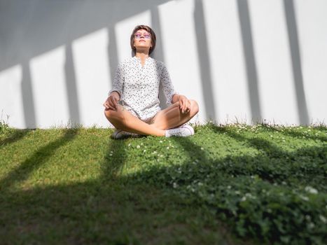 Woman is meditating on lawn in urban park. Nature in town. Relax outdoors after work. Summer vibes.