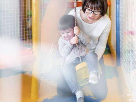 Toddler plays on rope swing with his mother or babysitter. Physical development for little children. Interior of kindergarten or nursery.