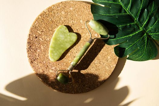 Jade Gua sha scraper and face roller massager on a cork round stand with a monstera leaf. Hard light, shadows, the concept self-care. Facial care. Zero waste. Lifting and toning treatment at home.
