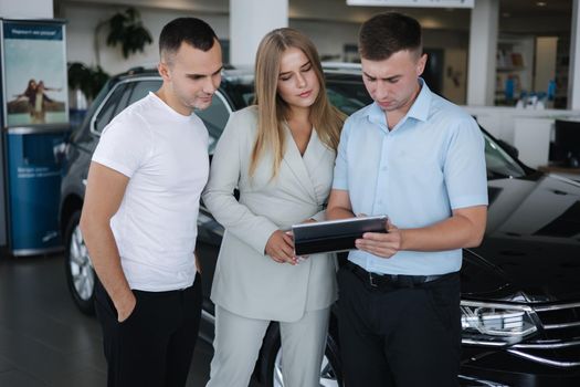 Salesperson talking with couple and help choosing new car. Man and woman in car showroom. Man using tablet to shop opportunity of different models.
