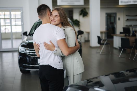 Gorgeous woman hug his husband after buying car in car showroom. Man and woman buy new car. Happy couple have new transport.