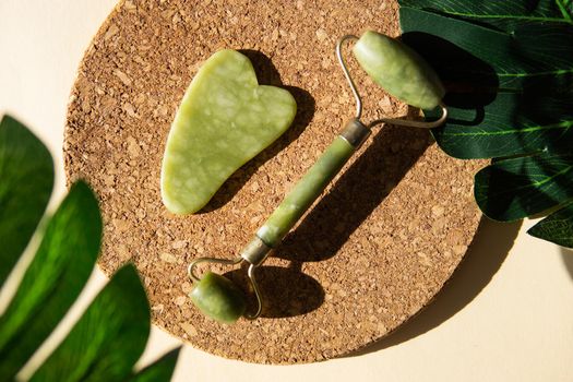 Jade Gua sha scraper and face roller massager on a cork round stand with a monstera leaf. Hard light, shadows, the concept self-care. Facial care. Zero waste. Lifting and toning treatment at home.