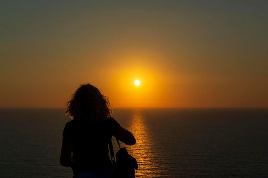 Silhouette of a woman at orange sunset on a cliff overlooking the sea at Palaiokastro castle of ancient Pylos. Greece.