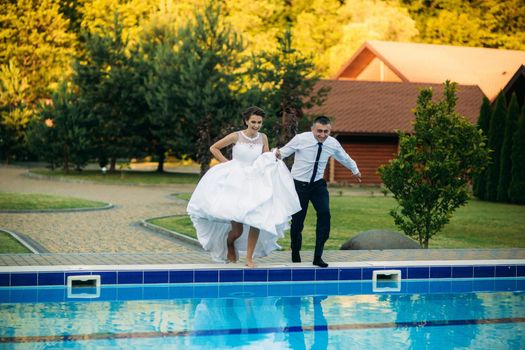 Young couple jumping in the swimming pool in a wedding suit and wedding dress. Sunny day.