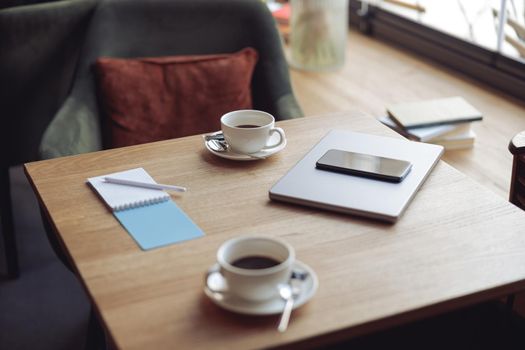 Wooden table with two white cups of coffee and business stuff. Laptop, notebook and phone. Blurred.