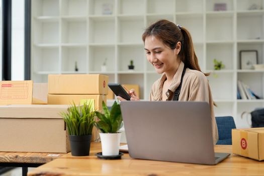 Young smiling beautiful owner asian woman freelance sme business online shopping working on laptop computer with parcel box on desk at home