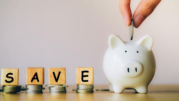 Hand of businessman is putting a coin into piggy bank near stack coins with the word save is written from a wooden block or cube. Loan, property ladder, financial, real estate investment concept.