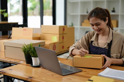 Young smiling beautiful owner asian woman freelance sme business online shopping working on laptop computer with parcel box on desk at home