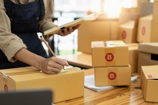 Young asian woman writing address on parcel box at home for delivery order to customer, shipping and logistic, merchant online and seller, business owner or SME, online shopping and e-commerce.