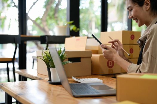 Young smiling beautiful owner asian woman freelance sme business online shopping working on laptop computer with parcel box on desk at home