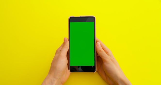 Woman watching content videos on Green Screen Chroma Key smartphone on yellow table background. Phone green screen for product placement. Creative background.