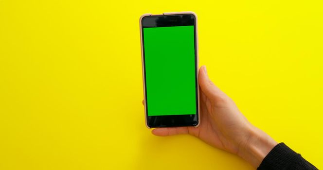 Woman watching content videos on smartphone with Green Mock-up Screen Chroma Key on yellow table background. Phone green screen for product placement. Creative background.