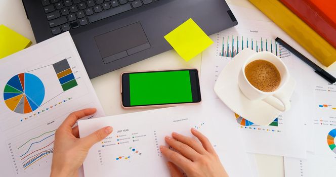 Top view of smartphone with green screen on the office desk. Application for business. Laptop, worksheet finance, graph accounts on office table workspace. Chroma key. Analysis of financial report