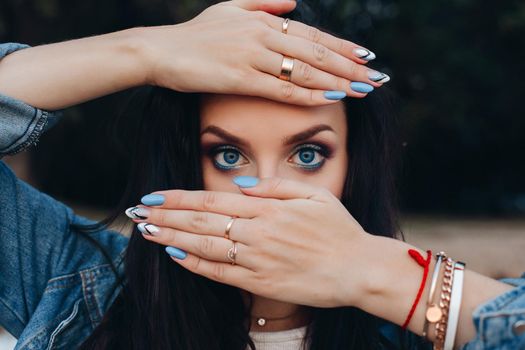 Beautiful woman with long dark hair wearing jewelry. Gorgeous girl with amazing make up in jeans jacket holding arms near head. Pretty young lady hiding behind hands with elegant blue and white nails.