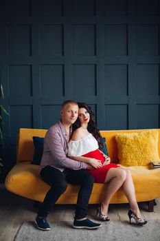 Family waiting for baby and loving each other. Elegant and stylish couple sitting on yellow couch and looking at camera. Blonde husband hugging pregnant brunette wife and holding hands on her belly.