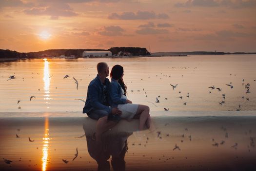 Romantic evening of family near picturesque bay. Side view of couple of young people in casual clothes sitting near water during sunset. Brunette woman and blonde man looking at birds and relaxing.