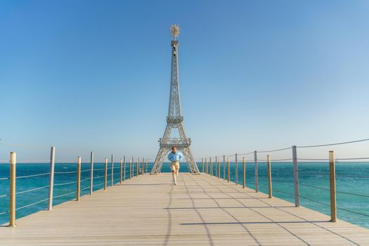 Large model of the Eiffel Tower on the beach. A woman walks along the pier towards the tower, wearing a blue jacket and white jeans