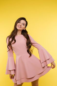 Beautiful young girl wearing short pink dress and trendy hairstyle looking at camera and posing on yellow isolated background. Attractive woman modeling in studio. Concept of fashion and beauty.
