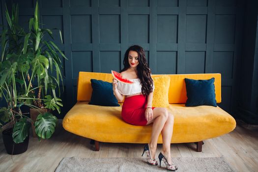 Stylish pregnant woman sitting on orange sofa, eating watermelon and posing. Attractive future mother in elegant clothes awaiting for little baby and relaxing. Concept of pregnancy and fashion.