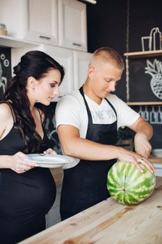 Happy young couple of people standing at kitchen and eating fruits. Beautiful brunette pregnant woman holding plate and standing near husband. Handsome man cutting watermelon and sharing it with wife.