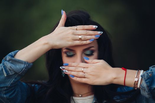 Front view of attractive brunette posing with closed eyes. Beautiful female with professional makeup and hairstyle standing in park. Young girl showing delicate manicure. Concept of beauty.