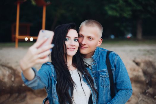 Lovely couple walking in park and taking selfie together. Happy girlfriend and boyfriend wearing jeans jackets dating and enjoying free time in evening. Concept of love and happiness.