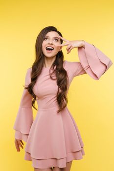 Excited stunning lady with long wavy hairstyle wearing trendy pink blouse showing ok gesture looking up with open mouth. Isolate on yellow.