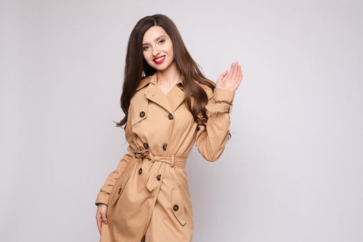 Brunette pretty lady with long hair in elegant coat posing at camera on grey studio background. Beautiful young woman with red lips holding hand up and waving. Gorgeous happy model in trench smiling.