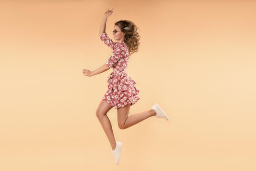 Side view of crazy girl wearing pink floral dress and white sneakers jumping and laughing in studio. Attractive young female in glasses enjoying warm weather and stylish outfit. Concept of happiness.