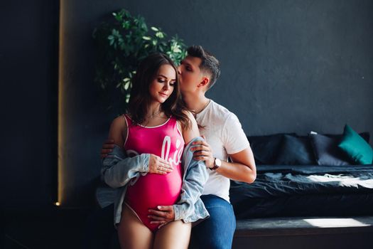 Portrait of attractive young couple sitting together. Beautiful pregnant woman and handsome man hugging and holding hands together on female belly. Happy family in casual clothes posing at camera.