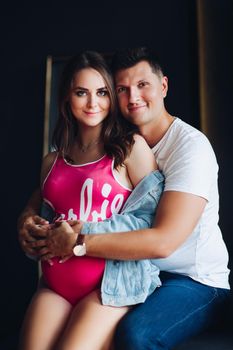 Portrait of attractive young couple sitting together. Beautiful pregnant woman and handsome man hugging and holding hands together on female belly. Happy family in casual clothes posing at camera.