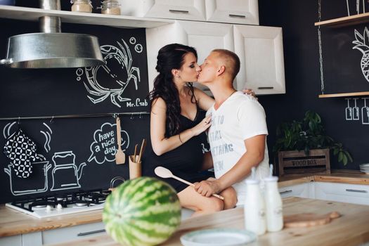 View from side of cute pair kissing while cooking at kitchen. Young pregnant woman expecting little baby with caring husband and resting together at home. Concept of pregnancy and love.