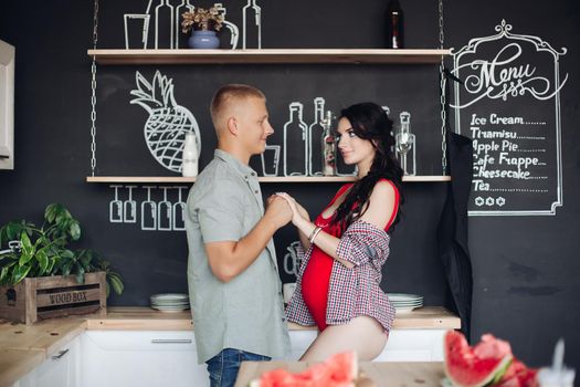 Happy family waiting for baby and loving each other. Pregnant brunette wife looking at her blonde handsome husband. Attractive couple of young people standing in kitchen and holding hands together.