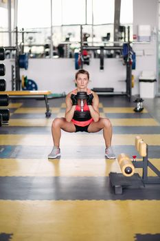 Sporty woman wearing stylish sport wears, making exercise squatting with dumbbells in gym. Athletic girl standing on special floor covering with simulators on background. Concept of healthy lifestyle.