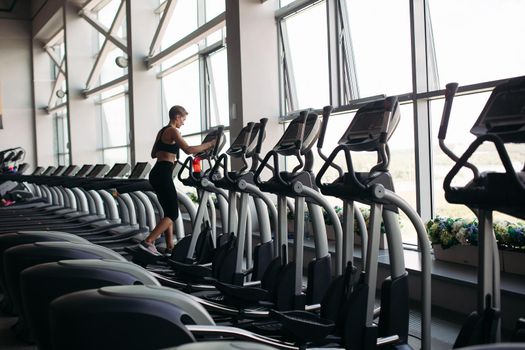 Young attractive fitness woman running on treadmill at panoramic window, wearing in black sportswear. Healthy sporty woman doing cardio exercise on treadmill. Concept of sport and healthy lifestyle.