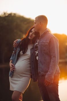 Beautiful pair expecting little child. Lovely parents standing near lake, hugging each other and posing. Pretty pregnant woman touching belly and enjoying motherhood. Concept of love and pregnancy.