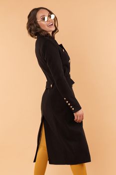 Side view of stylish girl in long black coat and elegant sunglasses on light studio background. Gorgeous happy model standing halfly turned and smiling. Brunette woman posing in fashionable clothes.