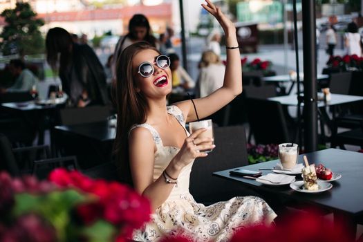 Portrait of a beautiful joyful brunette woman in a cafe. She is dressed in a sumptuous stylish pink dress and stylish glasses. He handsily lifts his hand up and holds a cocktail. Concept of a joyful woman in a cafe.